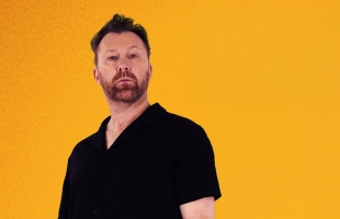 Jason Byrne stands staring towards the camera with a firm expression. He is wearing a black short sleeve shirt against an orange background.
