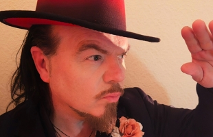 Jack L wearing a wide-brimmed hat with a red underside looks to the side, raising his hand slightly as if shading his eyes. He has a short beard and moustache, long hair, and wears a dark jacket with a pink rose on the lapel.