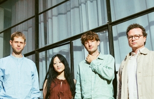 A outdoor group portrait of four people standing side-by-side in front of a modern glass-paneled building with sheer white curtains visible inside. From left to right: a man in a light blue collarless shirt, a woman with long dark hair in a rust-brown sleeveless top, a man in a green striped linen button-up holding his hand near his collar, and a man with glasses wearing a beige jacket over a light shirt.