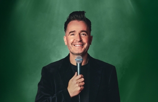 Jarlath Regan in a black suit stands holding a microphone up to his smiling face, with a green backdrop
