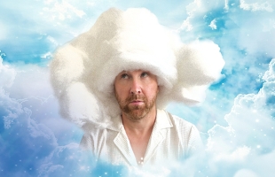 Jason Byrne, with a thoughtful expression, is shown against a dreamy sky filled with fluffy clouds. He wears a white shirt and has an oversized, cloud-like hat on his head, blending into the cloudy background.