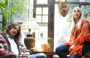 Paul Jorvas sitting cross-legged, head tilted, and Kathryn Pepper sitting opposite him. Both are dressed in flowing tops and jeans, and appear to be in a conservatory.