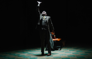 Man in a dark suit stands alone under a spotlight on a stage, reaching one hand upward dramatically beside an orange chair in a dark theatre.