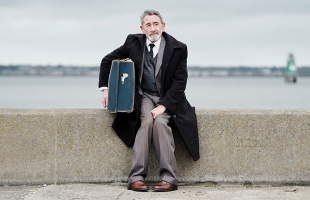 An older man in a dark overcoat sits on a concrete seawall, holding a worn suitcase, with a calm body of water and cloudy sky behind him.