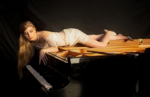 Kelly Moran in a white dress and shoes, lying across a grand piano. Some of the piano keys are depressed, suggesting that it is playing by itself.