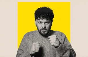 A black-and-white portrait of a Killian Sundermann - a  man with curly dark hair and a short beard - wearing a thick knit jumper, holding his fists up in a playful boxing stance against a bright yellow background.