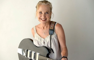 Kristin Hersh stands centre shot, she is against a grey background, she is standing toward the camera smiling, she is holding her guitar which is hanging off her shoulder.