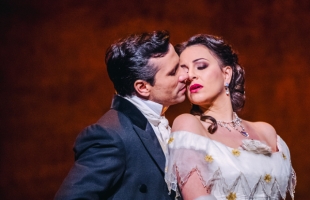 Two opera performers in period costumes share an intimate moment onstage. The man, dressed in a dark formal suit with a white cravat, leans in as if to kiss the woman’s cheek. The woman, wearing an elegant off-the-shoulder white gown with gold accents and ornate jewelry, tilts her head back with a dramatic, emotional expression. Warm, theatrical lighting highlights their faces against a blurred background.