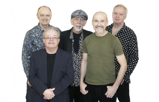 Five members of Lindisfarne stand together against a white background, they are looking towards the camera smiling, Rod Clements stands in the centre wearing a cap, shits and blazer.