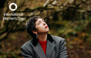 Lisa O'Neill wearing a grey blazer and red jumper, looking wistfully to the sky, with a forest behind her.