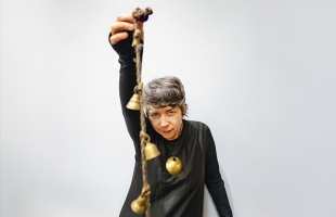 Lisa O'Neill in a long-sleeve grey top, with her right arm stretched above her head, holding a rope with small golden bells hanging off it.
