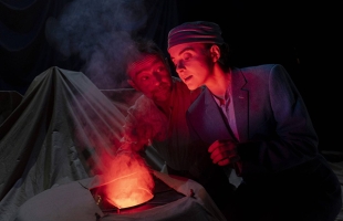 Dramatic shot of two actors an older man and younger women, huddled around what looks like a fire.