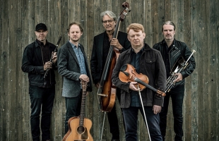 Five men stand against a weathered wooden wall, holding traditional instruments including fiddle, flute, uilleann pipes, guitar and double bass, dressed in dark, casual clothing.
