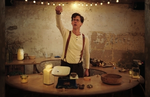 Manchán Magan at a weighing scales gesticulating as if explaining something. He wears suspenders and a white, longsleeve t-shirt.
