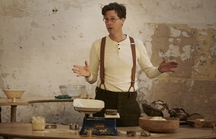 Manchán Magan at a weighing scales gesticulating as if explaining something. He wears suspenders and a white, longsleeve t-shirt.