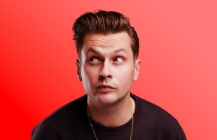 Headshot portrait of Mark Mehigan set against a vibrant solid red background. He is looking upward and off to the side with an expressive, quizzical look, styled with swept-back brown hair, wearing a dark sweater and a thin gold chain.