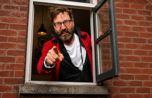 McSavage, a middle-aged man with wild, messy dark hair, glasses, and a full beard, leans out of an open ground-floor window set in a red-brick wall. He makes an exaggerated, intensely angry face while pointing directly forward at the camera. He wears a white t-shirt under a black hoodie and a bright red jacket.