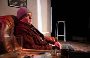 Person in a knitted hat and chunky cardigan sits in a leather armchair on a living-room stage set, gazing thoughtfully into the distance.