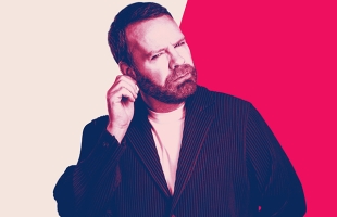 Neil Delamere with a puzzled expression scratches his ear, appearing to listen closely or think through a question.