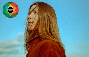Niamh Regan stands with a blue sky behind her, wearing a red turtle neck, and her red hair windswept across her face