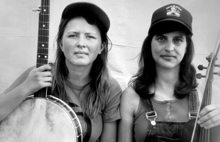 Nora Brown holding her banjo, and Stephanie Coleman with her fiddle, smiling in black and white