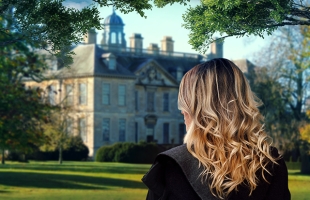A woman with long wavy blonde hair stands in a sunlit garden, looking toward a large historic mansion framed by trees.