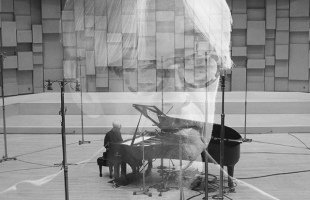 An faint, close, black-and-white headshot of Ryuchi Sakamoto wearing glasses, superimposed over a wide shot of Sakamoto sat in a large studio playing a grand piano.
