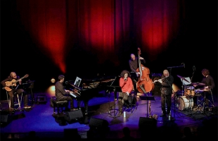 A live musical ensemble performing on a stage illuminated by warm red spotlighting against a dark background. The band includes a seated classical guitarist on the far left, a pianist at a grand piano, a vocalist in an orange top at center mic, an upright bassist behind her, a flutist, and a drummer on the far right.