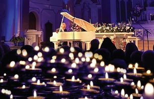 Phil Coulter playing a white grand piano in front of a silhouetted audience, and a sea of candles