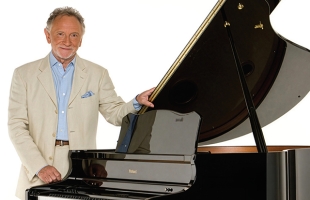 Phil Coulter in a cream blazer and light blue shirt stands beside a glossy black grand piano with the lid open, smiling at the camera.