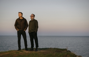 Paul Noonan and Brian Crosby stand on a cliff's edge with the sea behind them