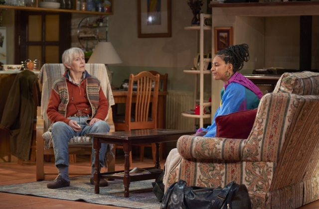Jane Brennan, dressed in a red fleece and jeans, sits opposite Fanta Barrie in a multicoloured wind breaker, a large duffle bag to the side of the couch