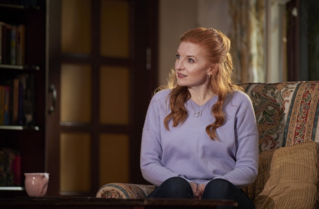Mags McAuliffe sits on a patterned couch, wearing a purple jumper, looking concerned