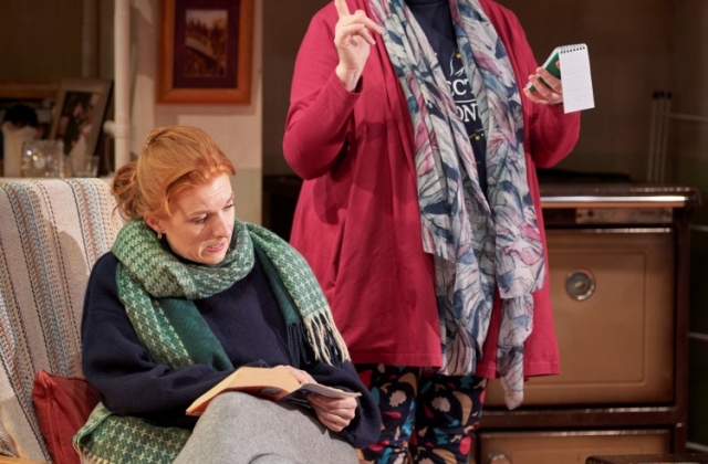 Mags McAuliffe, dressed in grey sweatpants, a navy jumper, and an oversized green scarf, sits on a chair reading a book while Helen Norton in a pink cardigan and patterned leggings stands holding a small notebook