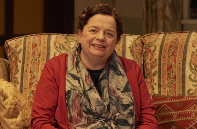 Helen Norton, smiling, sitting on a patterned couch. She is wearing a red top with a floral scarf.