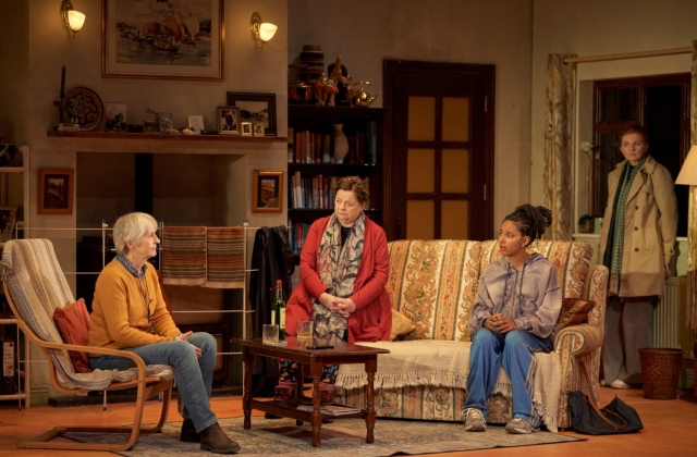 Jane Brennan sits in a chair, with Helen Norton and Fanta Barrie on a couch in a living room, while Mags McAuliffe looks on from stage right