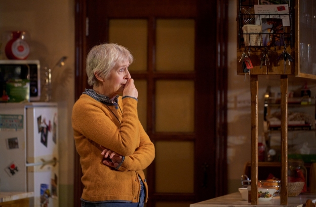 Jane Brennan, wearing a yellow cardigan, stands in a kitchen set, looking upset with her hand to her mouth