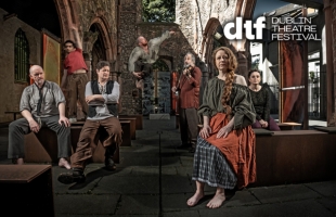 Seven performers are posed inside the roofless stone ruins of a church or abbey, surrounded by Gothic arches and weathered walls. A barefoot woman with long red hair sits prominently in the foreground wearing layered period-style clothing in rust, grey and olive tones. Other cast members are seated, standing or leaning throughout the space, creating a dramatic tableau. The scene is lit with theatrical contrast, highlighting the historic architecture. The Dublin Theatre Festival logo appears in the corner.