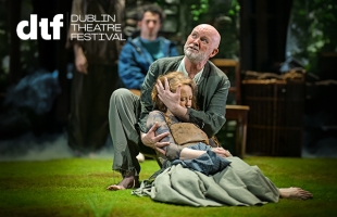 Performer kneels on a grassy stage, supporting another performer lying wrapped in layered, earth-toned clothing and a blanket. The kneeling performer cradles the other’s head. A third performer and wooden set pieces appear blurred in the background with soft, misty lighting. White text in the top left reads “dtf” and “Dublin Theatre Festival.”