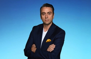 The image shows a man posing with his arms crossed against a solid blue background. He is wearing a dark blue blazer over a plain white t-shirt. A bright yellow pocket square peeks out from his breast pocket. He has dark, short hair styled back and a serious expression.