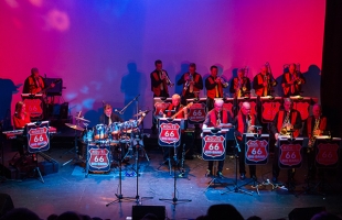 A live performance by the Route 66 Big Band on stage. The band members, dressed in coordinated black and red outfits, play various instruments including drums, keyboards, saxophones, trumpets, and trombones. Each music stand displays the Route 66 logo. The stage is lit with vibrant red and blue lighting, creating a dynamic and colourful atmosphere.