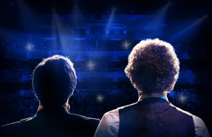 Two performers dressed as Simon and Garfunkel with their backs to the camera, staring out to a packed theatre.