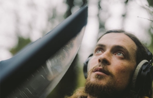 Seán Ronyane surrounded by forest, holds recording equipment up, listening on headphones