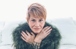 Shawn Colvin, a woman with short blonde hair, resting her crossed hands near her face, wearing a dark green fur wrap and showing a tattoo on her wrist against a pale background.