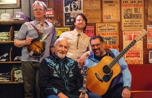 Members of Special Consensus gather with their instruments for a group portrait in a rustic bluegrass venue surrounded by vintage performance posters.
