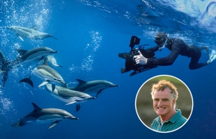 Ken O'Sullivan swims alongside a pod of dolphins in clear blue ocean water while filming with a professional camera rig. A circular inset in the corner shows him smiling outdoors.