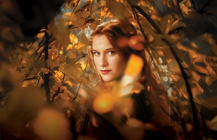 A redheaded young woman looks straight at the camera surrounded by orange, brown and red foliage.