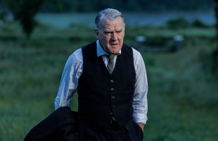 Mikel Murfi walks through a rural field, dressed in a suit, with the suit jacket over his right forearm, hands in his pockets