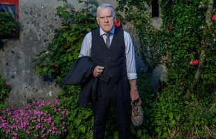 Mikel Murfi stands hunched in the garden of a rural cottage, dressed in a suit, and holding a battered shoe