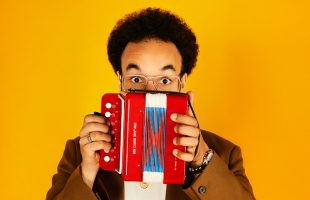 A close up shot of Théo Ould, he is pictured against a bright yellow background, holding up a small bright red and blue mini accordion, which covers the lower part of his face. He is wearing a glasses and he his eyes are open wide giving a surprised expression.